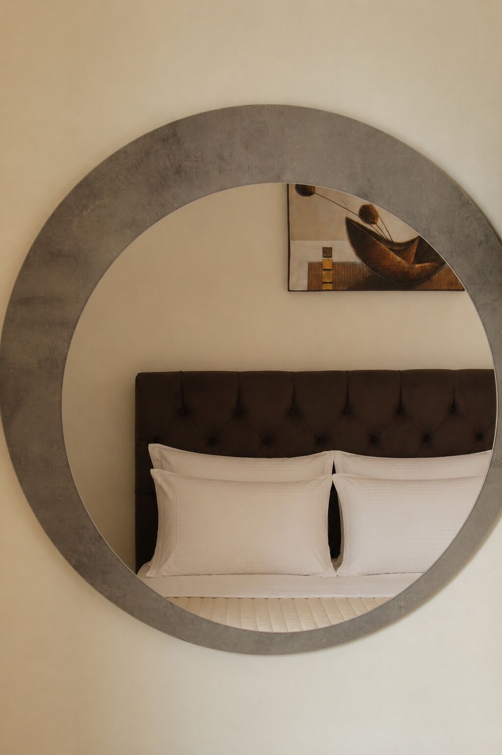 Una cama con cabecera capitonada, un espejo circular y un cuadro en la pared.