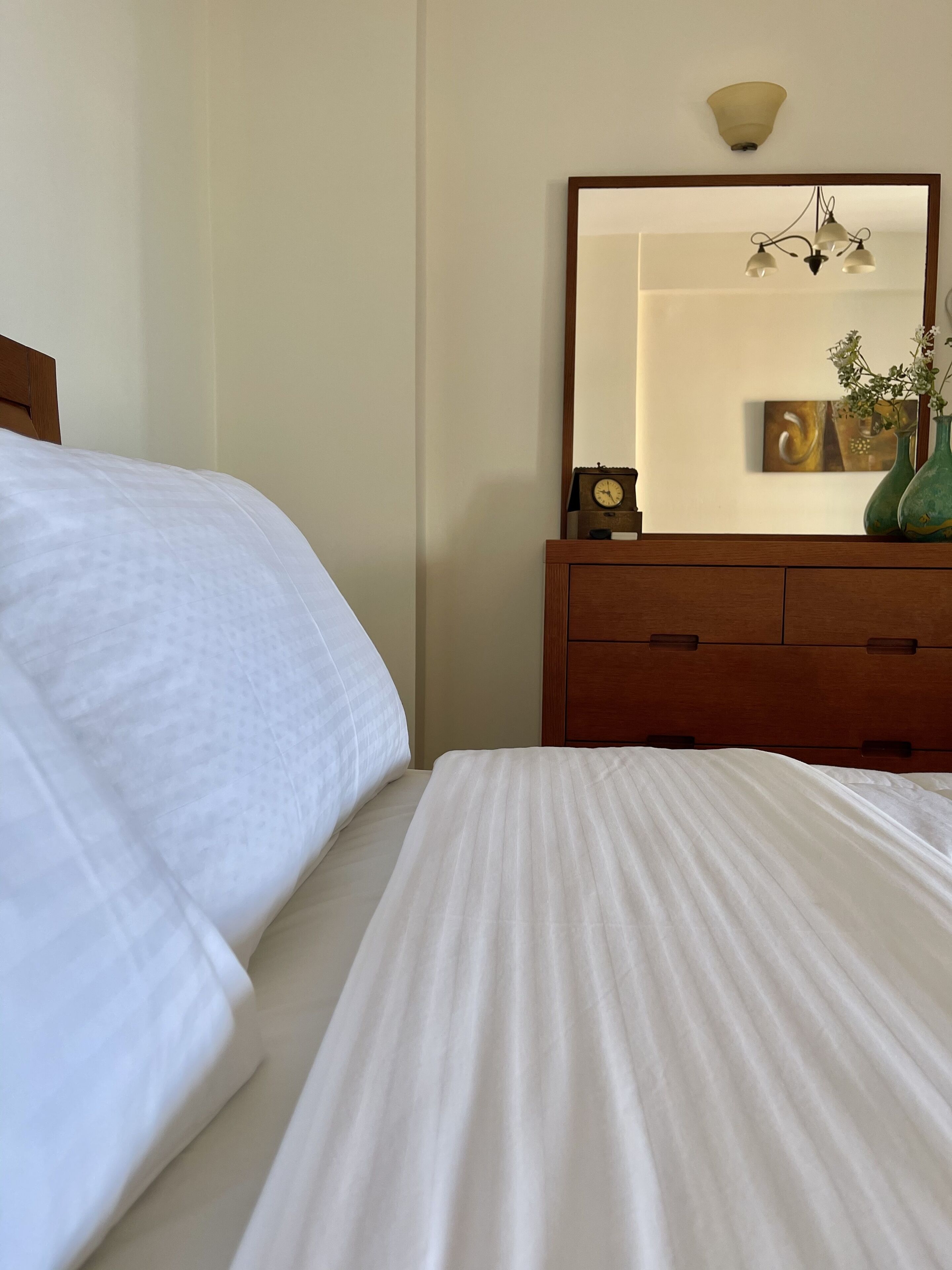A neatly made bed with white linens, a wooden dresser with a mirror above it, and a small clock on the dresser.