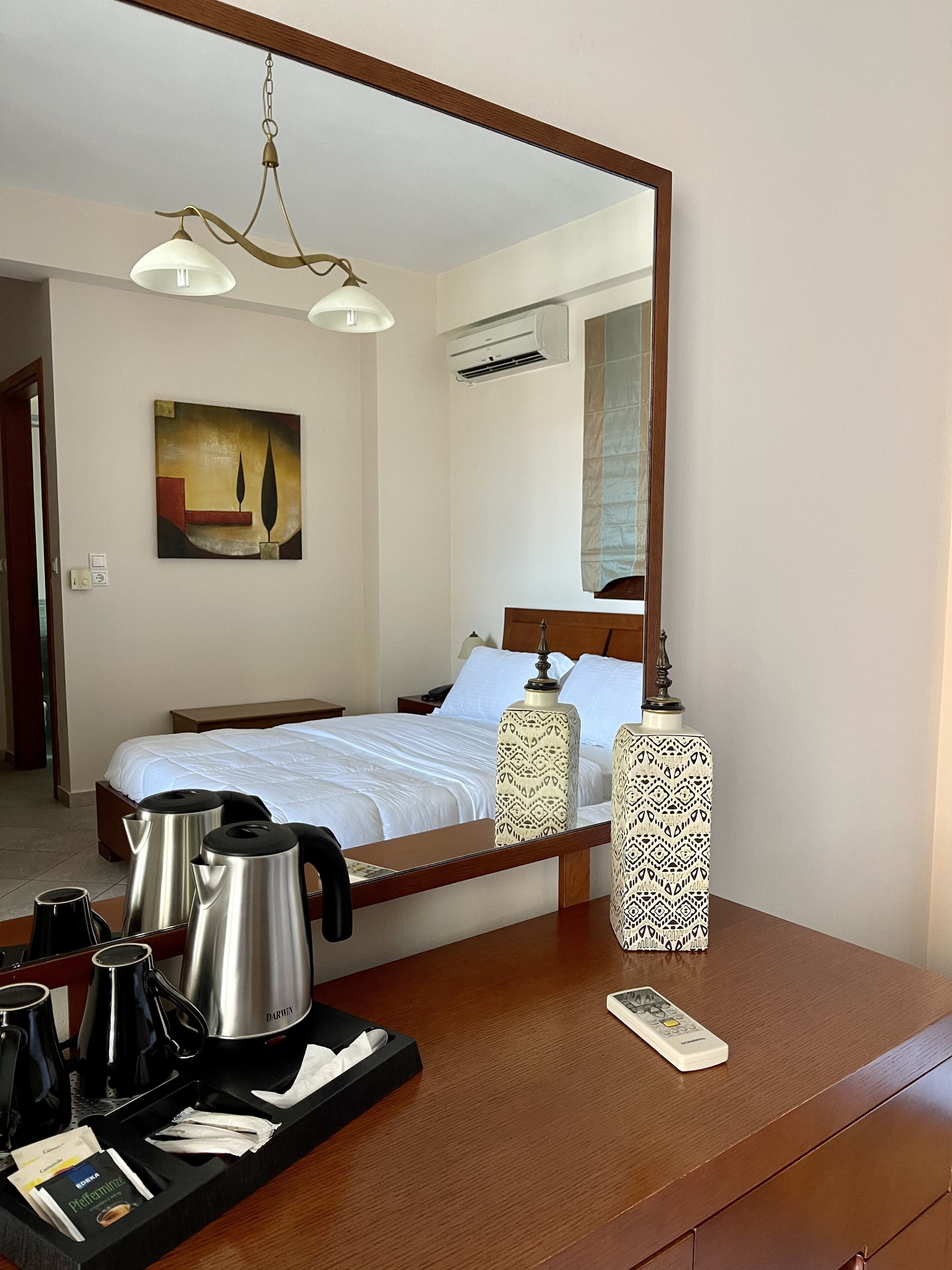 A hotel room with a bed, bedside table, coffee maker, remote control, and a mirror.