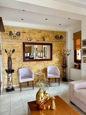 A living room with a stone wall, two white armchairs, a coffee table with decorative vases, a mirror, and a fireplace. - Sofotel (Pylos-Nestoras)