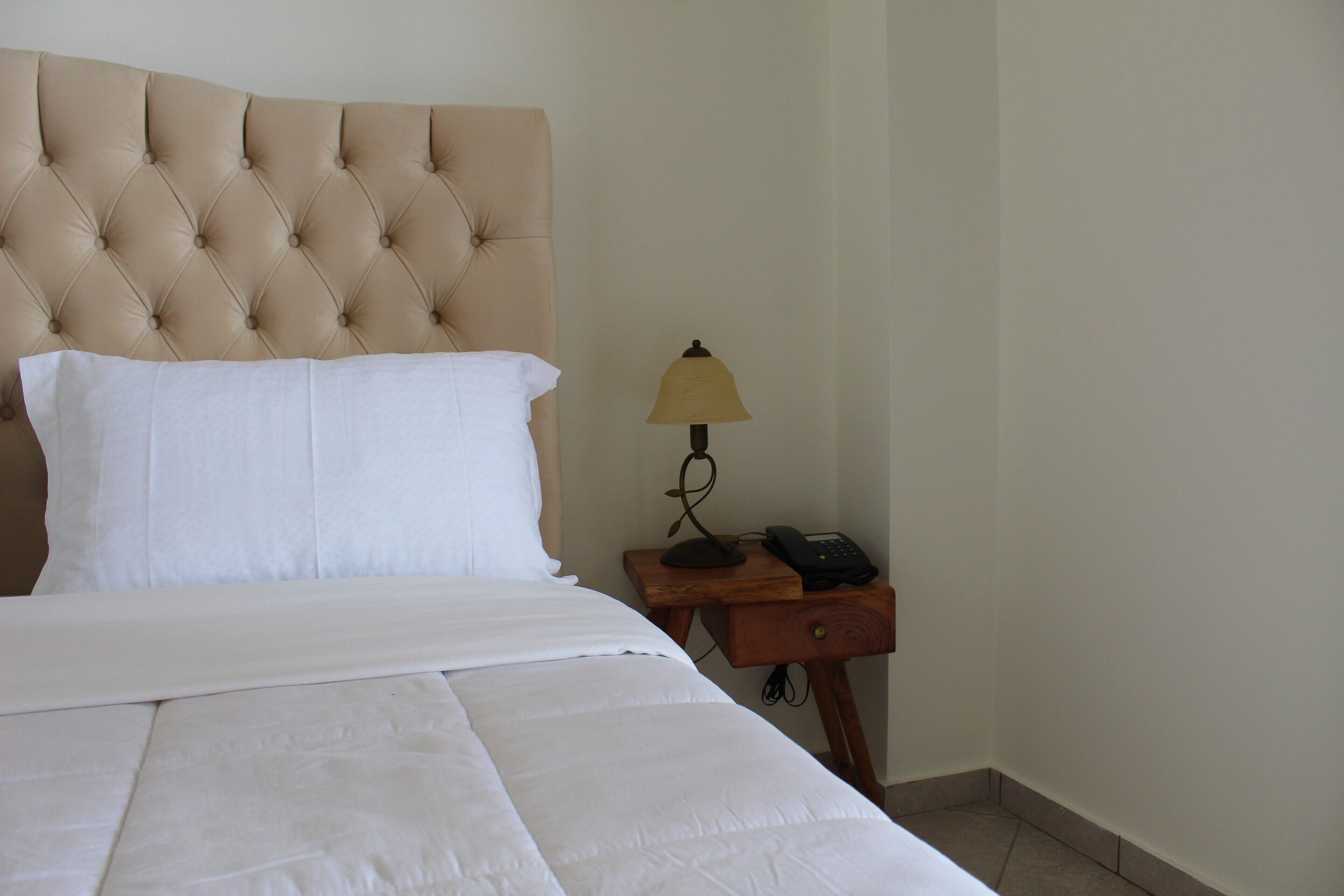 A neatly made bed with a tufted headboard, a bedside lamp, and a small wooden nightstand with a phone.