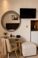Habitación moderna con espejo redondo, televisor de pantalla plana, escritorio con silla, pequeño otoman y área de kitchenette.