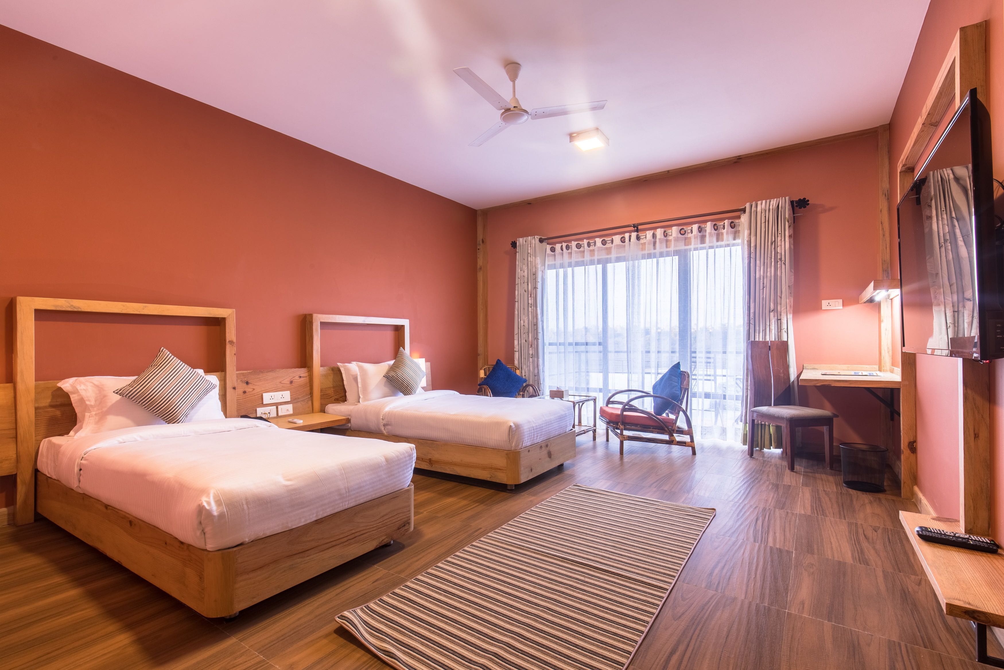 Quarto de hotel com duas camas, uma escrivaninha, uma cadeira, televisão e uma janela grande com cortinas.