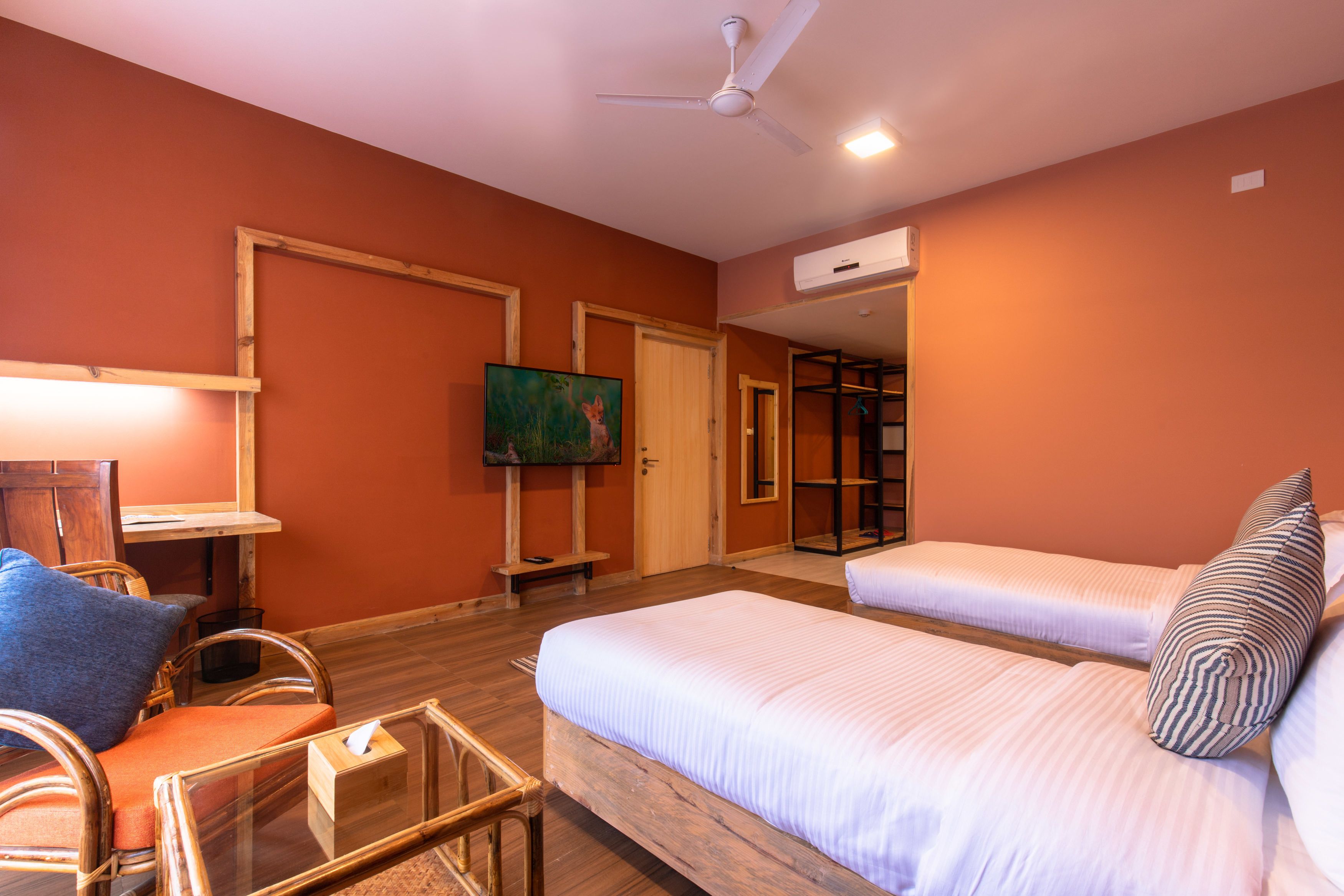 Une chambre d’hôtel avec deux lits, une chaise en bois, un bureau avec une lampe, une télévision et un ventilateur de plafond.