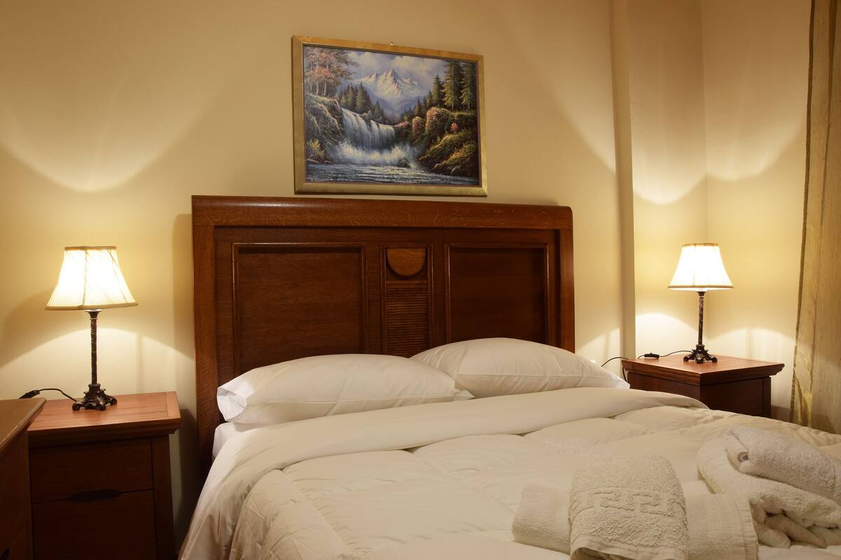 Uma cama bem arrumada com lençóis brancos e um cobertor, com duas mesas de cabeceira com luminárias, uma cabeceira de madeira e um quadro de uma cascata acima da cama.