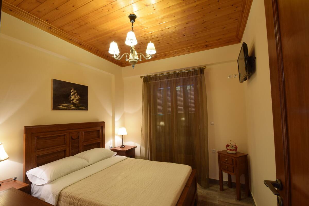 Quarto com teto de madeira, cama com roupa de cama branca, mesa de cabeceira com abajur, pequena mesa com um objeto decorativo e janela com cortinas.