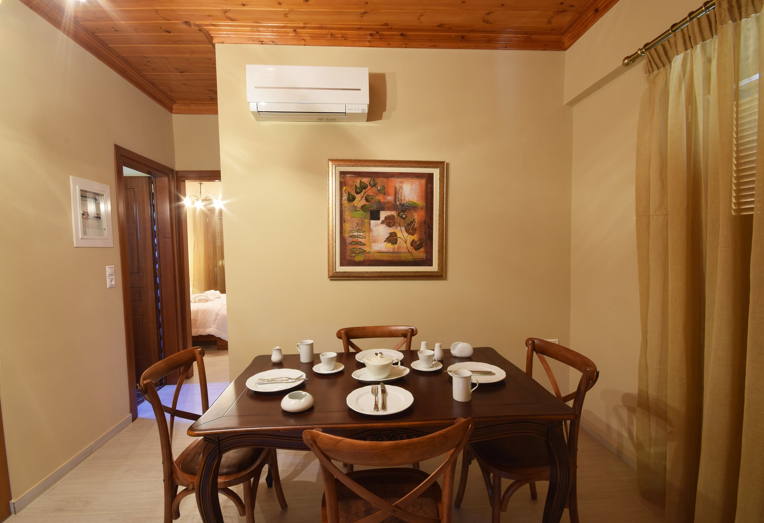 A dining room with a wooden table set for four, surrounded by wooden chairs. An air conditioning unit is mounted on the wall above the table.