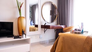 A hotel room with a flat-screen TV, a large vase with plants, a round mirror, a bedside table with toiletries, and a bed with a brown cover.