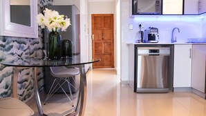 A modern kitchen with a stainless steel dishwasher, a microwave, and a coffee maker. There is a glass table with a vase of flowers and two chairs.