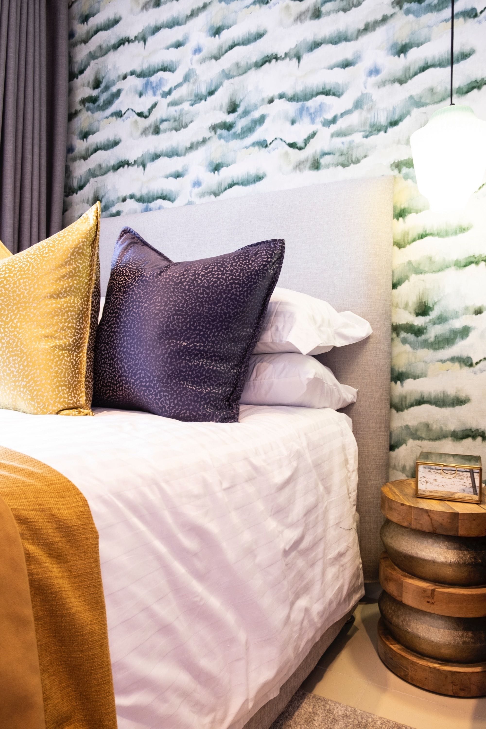 Una cama bien tendida con ropa de cama blanca y dorada, un papel tapiz estampado en la pared detrás y una mesita de madera con un cuadro enmarcado.