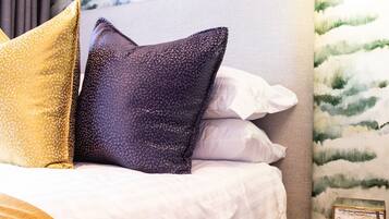 Una cama bien tendida con ropa de cama blanca y dorada, un papel tapiz estampado en la pared detrás y una mesita de madera con un cuadro enmarcado.