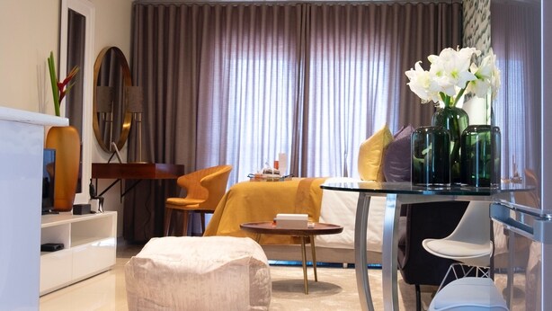 A modern living room with a glass dining table, comfortable seating, a sofa, and a large window with curtains.