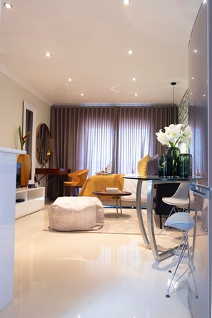 Una sala de estar moderna con una mesa de comedor de vidrio, asientos cómodos, un sofá y un ventanal con cortinas.