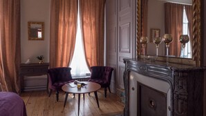 Una habitación con piso de madera, dos sillones morados, una mesa redonda con un vaso, una chimenea y un espejo grande que refleja tres copas de vino.