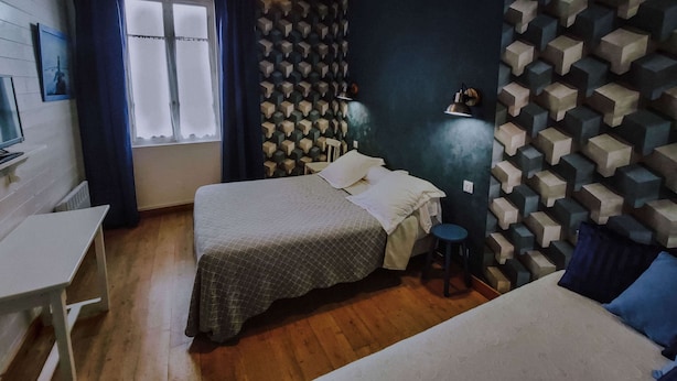 A room with a bed, a desk, a chair, a window with curtains, and a patterned wall. - Hôtel Le Marronnier (Tramayes)