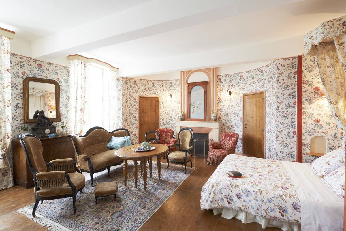 A traditional room with floral wallpaper, a fireplace, a bed, and a dining area with chairs and a table.