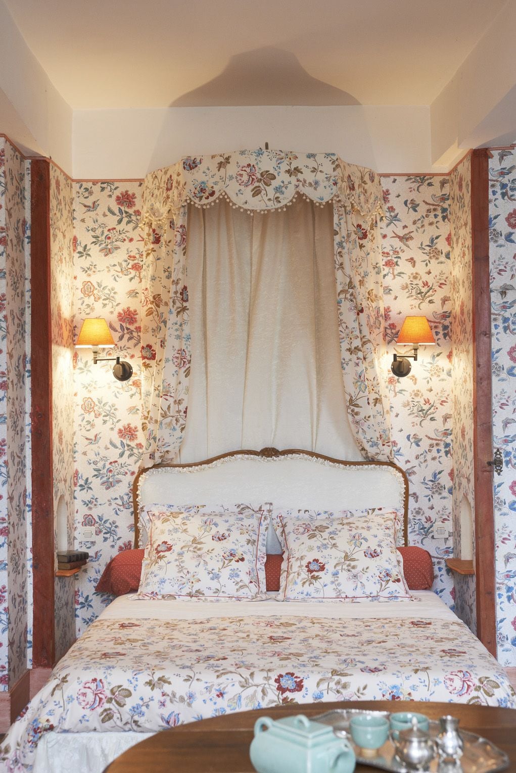 Une chambre à coucher avec un papier peint à motifs floraux, un lit à baldaquin, deux lampes fixées au mur et une table de chevet sur laquelle se trouvent une théière et des tasses.