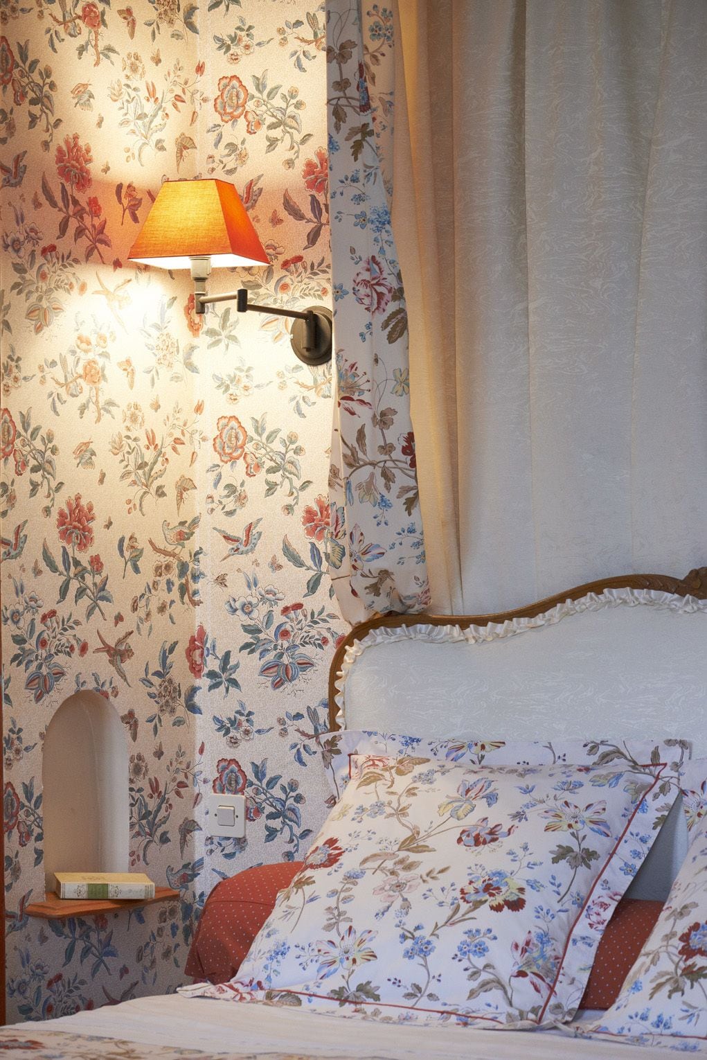 Un dormitorio con papel tapiz floral, una lámpara de noche, un cabecero de madera y una mesita de noche con un libro.
