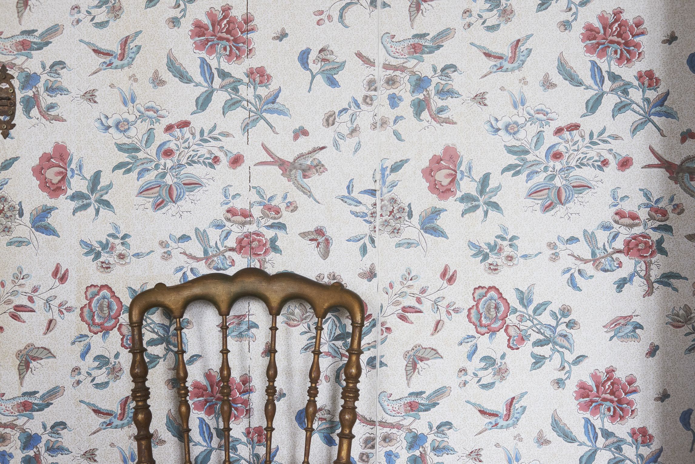 A chair with a wooden backrest and turned spindles against a floral wallpaper with birds and flowers.