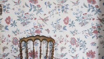 A chair with a wooden backrest and turned spindles against a floral wallpaper with birds and flowers.
