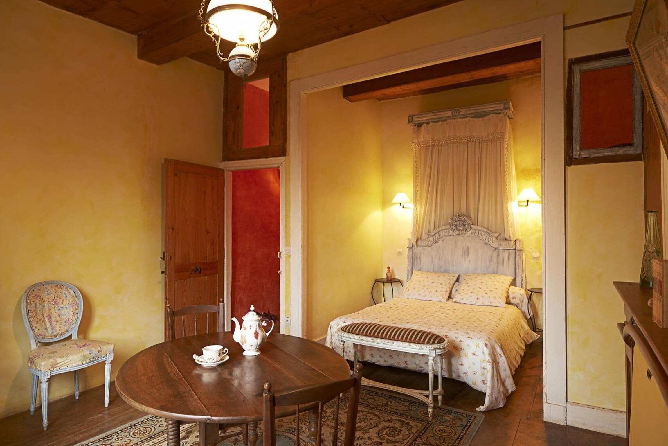 A room with a wooden dining table, a chair, a bed with a canopy, and a red door.