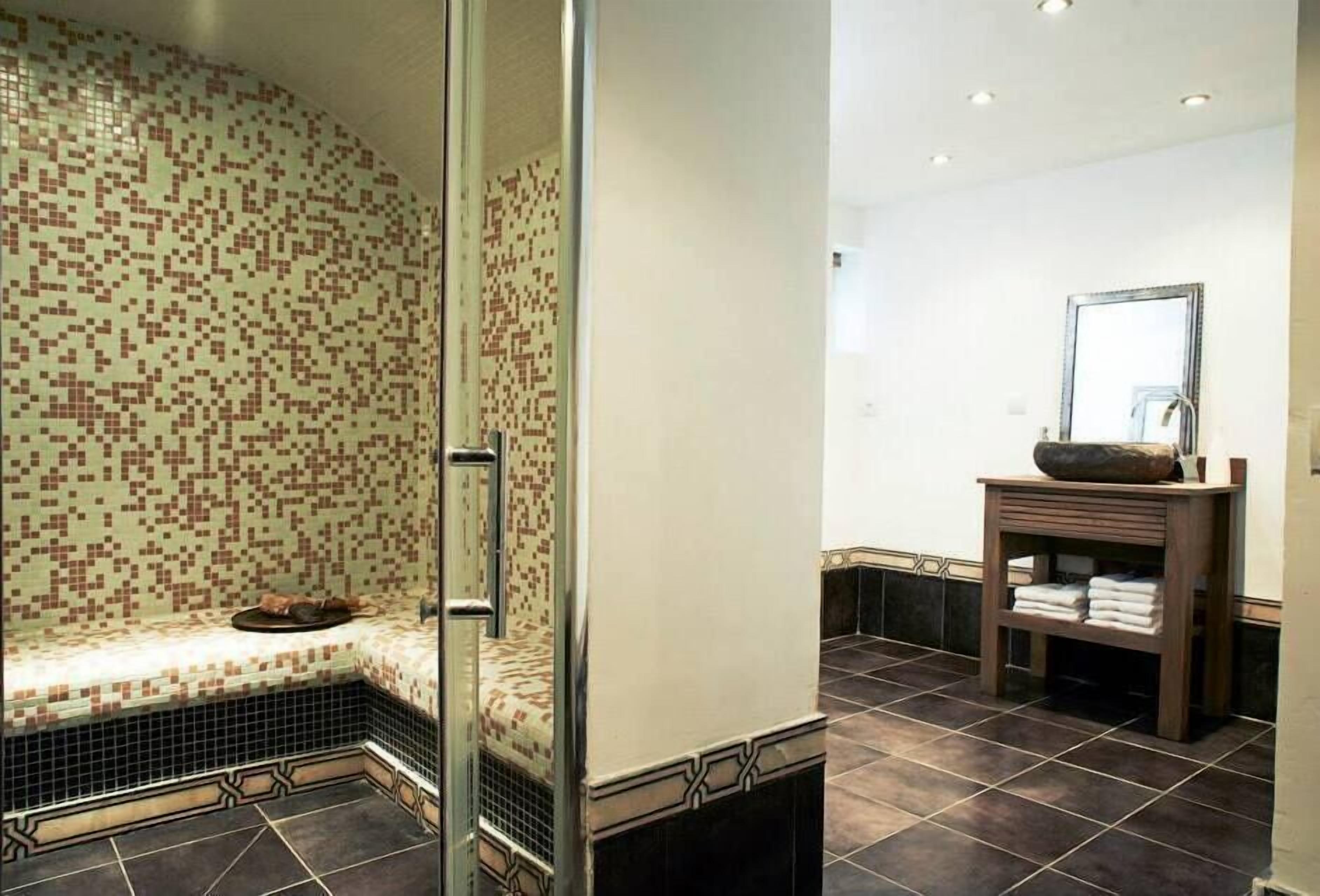 Une salle de bain avec un mur recouvert de mosaïques, une cabine de douche en verre, un meuble-lavabo en bois avec un miroir et un sol carrelé.