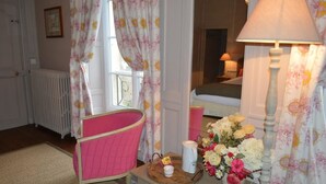 Quarto com uma cadeira rosa, uma mesa de cabeceira com abajur e flores, e uma janela com cortinas florais.