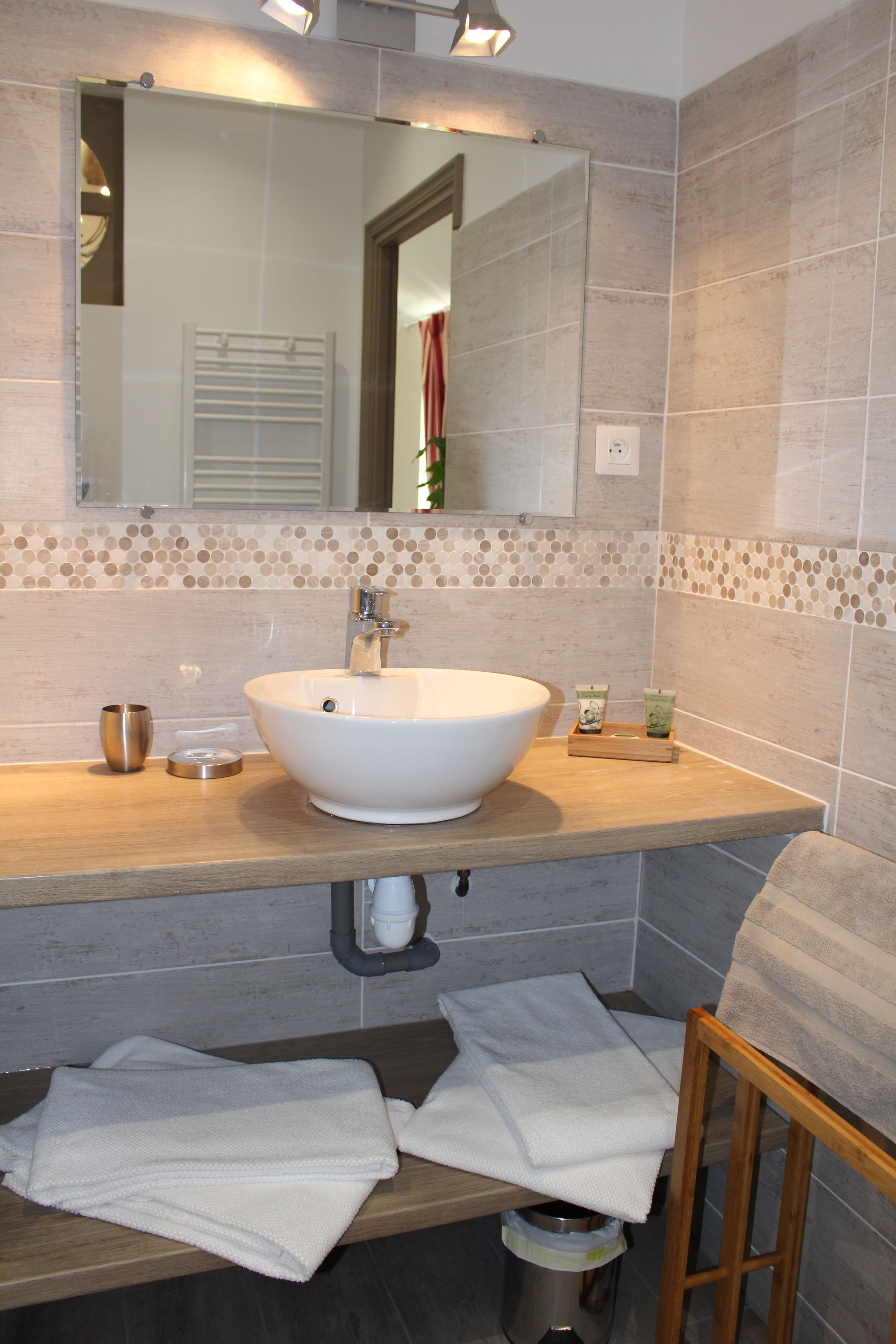 A bathroom with a white sink, mirror, and tiled walls. There are towels on the counter and a trash can below.