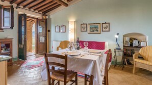 Una zona de menjador amb una taula preparada per a dues persones, una cadira de vimet, una petita taula auxiliar amb una làmpada i una biblioteca amb llibres i una cafetera.