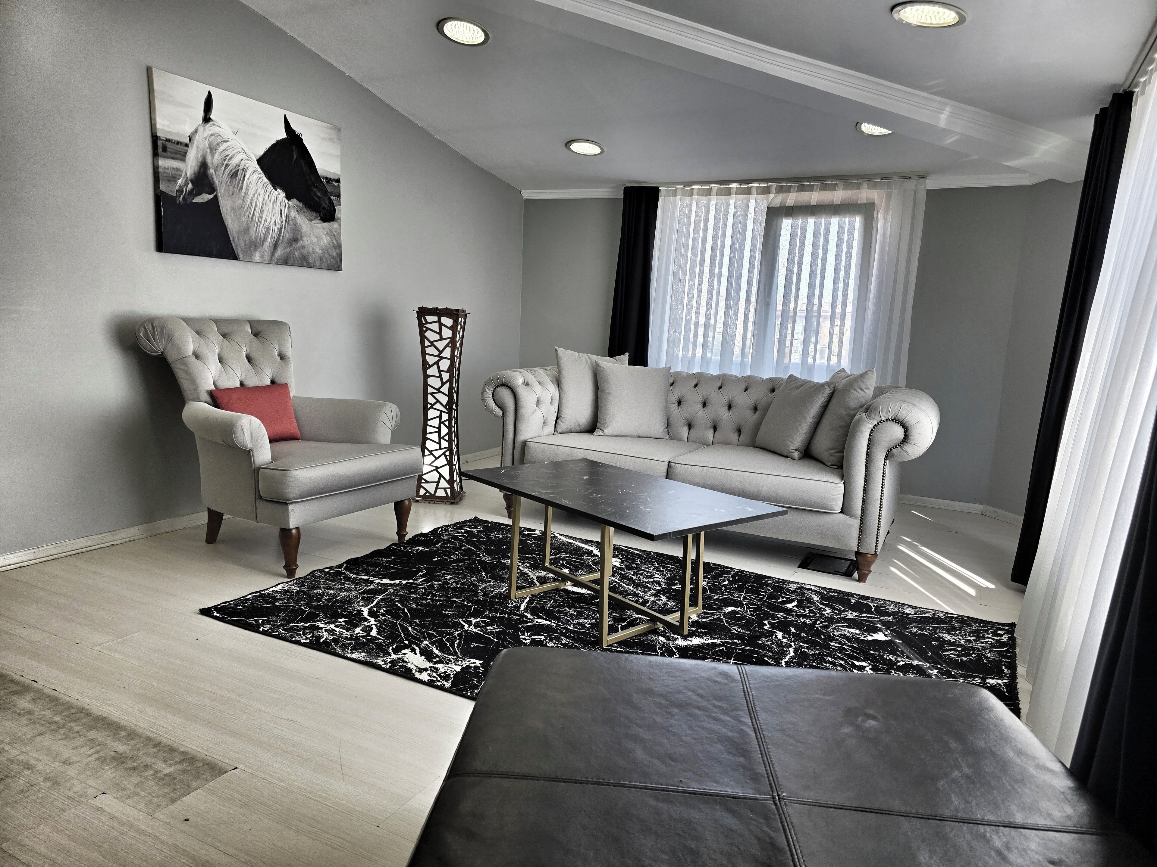 Un salon moderne avec un canapé gris, un fauteuil à capitons, une table basse noire et un pouf noir.