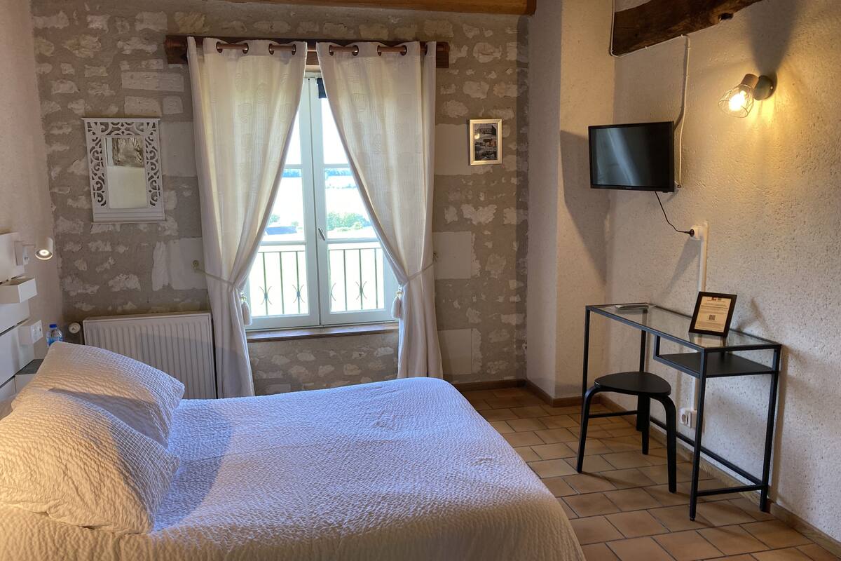 Ein Schlafzimmer mit Bett, einem Fenster mit Vorhängen, einer Heizung, einem kleinen Tisch mit Stuhl, einem an der Wand montierten Fernseher und einem Bilderrahmen.