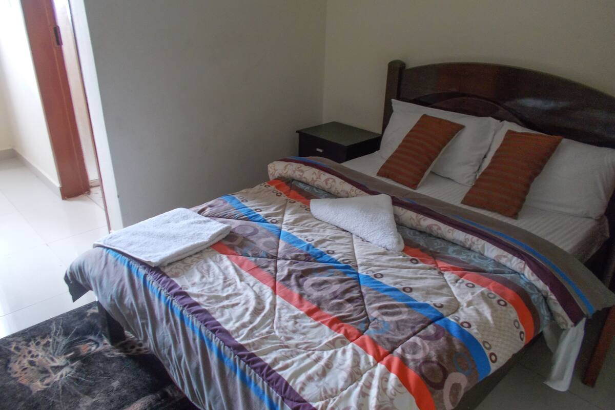 A neatly made bed with a patterned bedspread and pillows, a bedside table, and a window with a view of the outside.