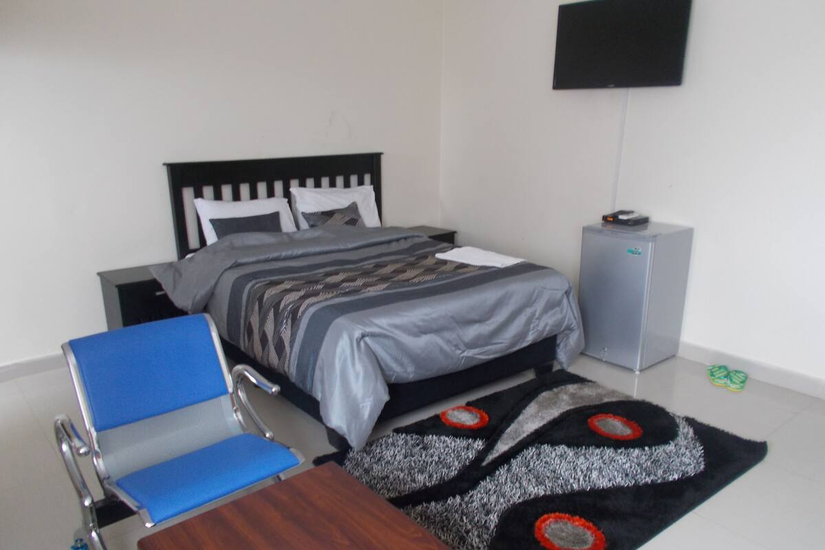 A bedroom with a bed, a desk, a chair, a small cabinet, and a TV on the wall.