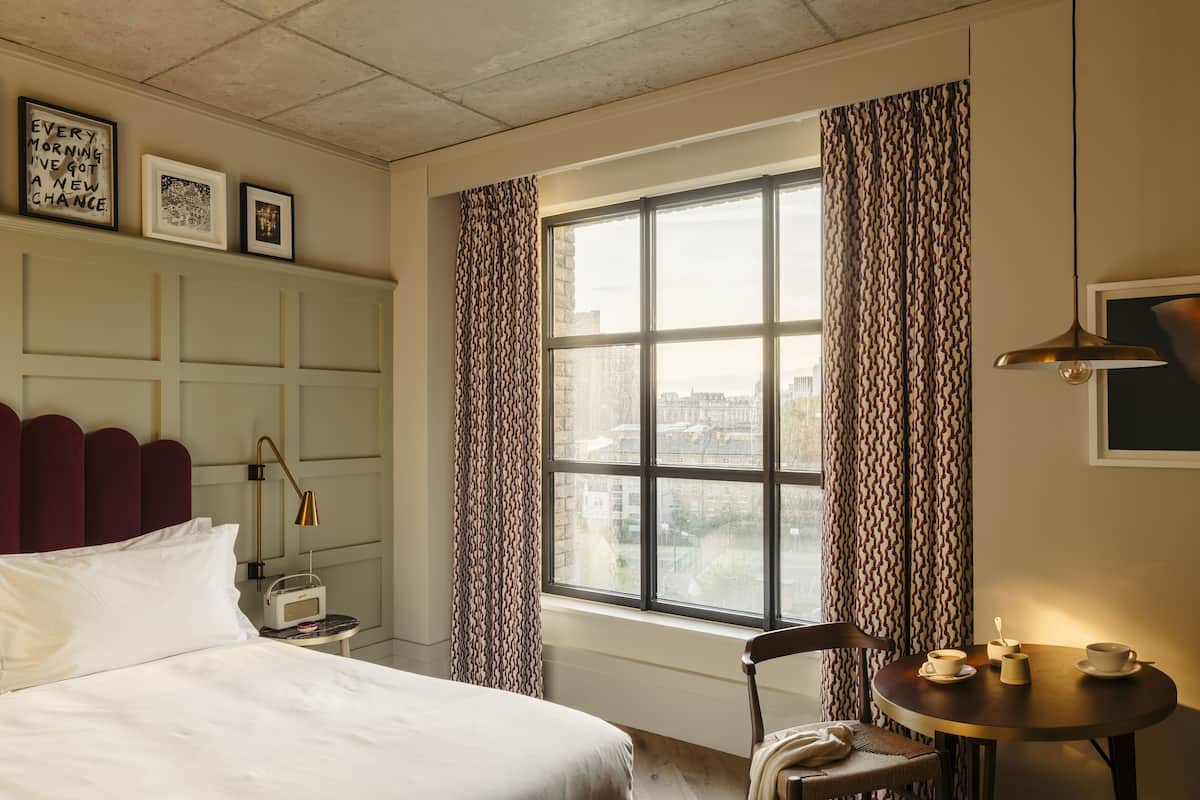 Ein Hotelzimmer mit großem Fenster, einem Bett, einem Nachttisch mit Radio, einem Sessel und einem kleinen Tisch mit Tassen und einem Löffel.