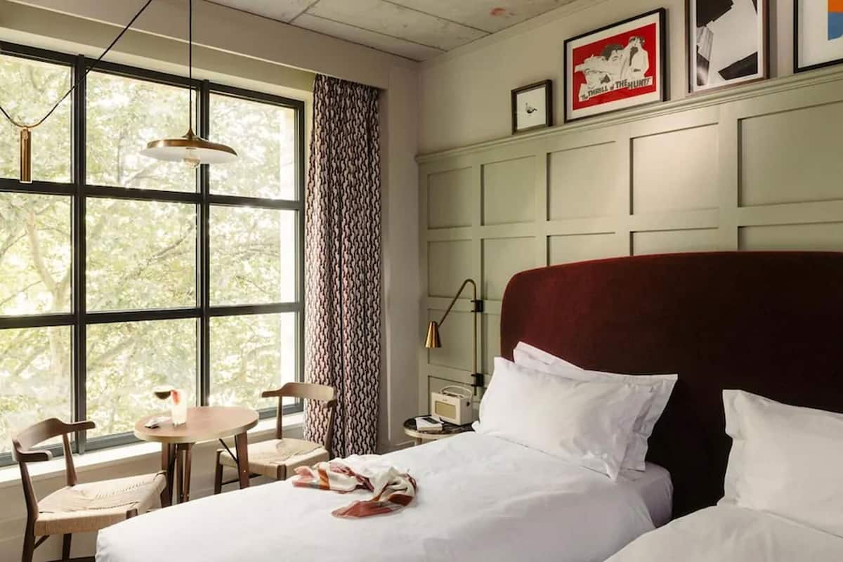 Ein Hotelzimmer mit einem großen Bett, einem kleinen Tisch, zwei Stühlen, einem Fenster mit Blick auf Bäume und einer Wand mit gerahmten Bildern.