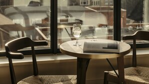 A table with a book and a glass of water, two chairs, and a pendant light, with a city view through large windows. - The Hoxton, Southwark (London)