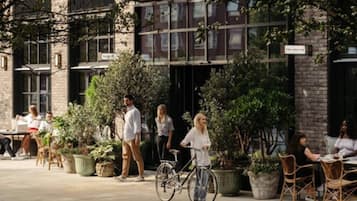 An outdoor seating area with tables and chairs, surrounded by potted plants and a building with large windows.