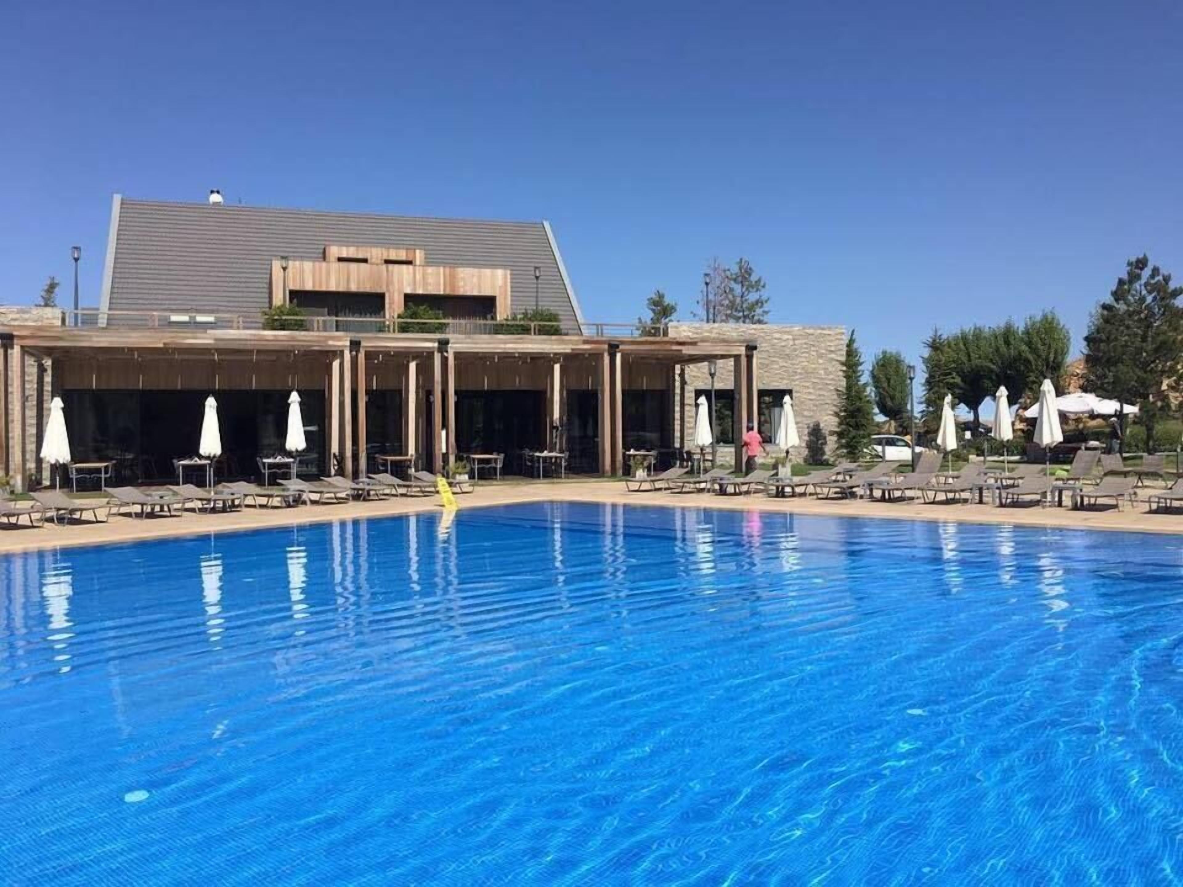 Una piscina con sillas de descanso y sombrillas, un edificio de fachada de piedra, y un cielo azul despejado.