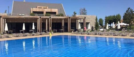 Una piscina con sillas de descanso y sombrillas, un edificio de fachada de piedra, y un cielo azul despejado.