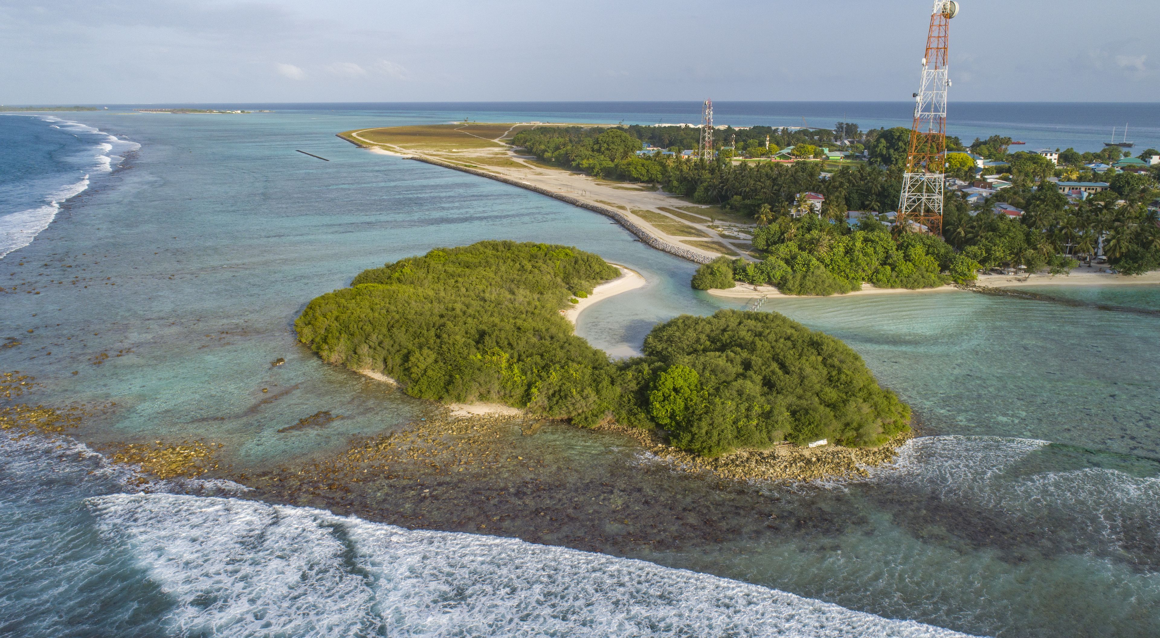 An aerial view of a coastal area with clear waters, two small islands with dense vegetation, and a distant shoreline with buildings and a tall communication tower.