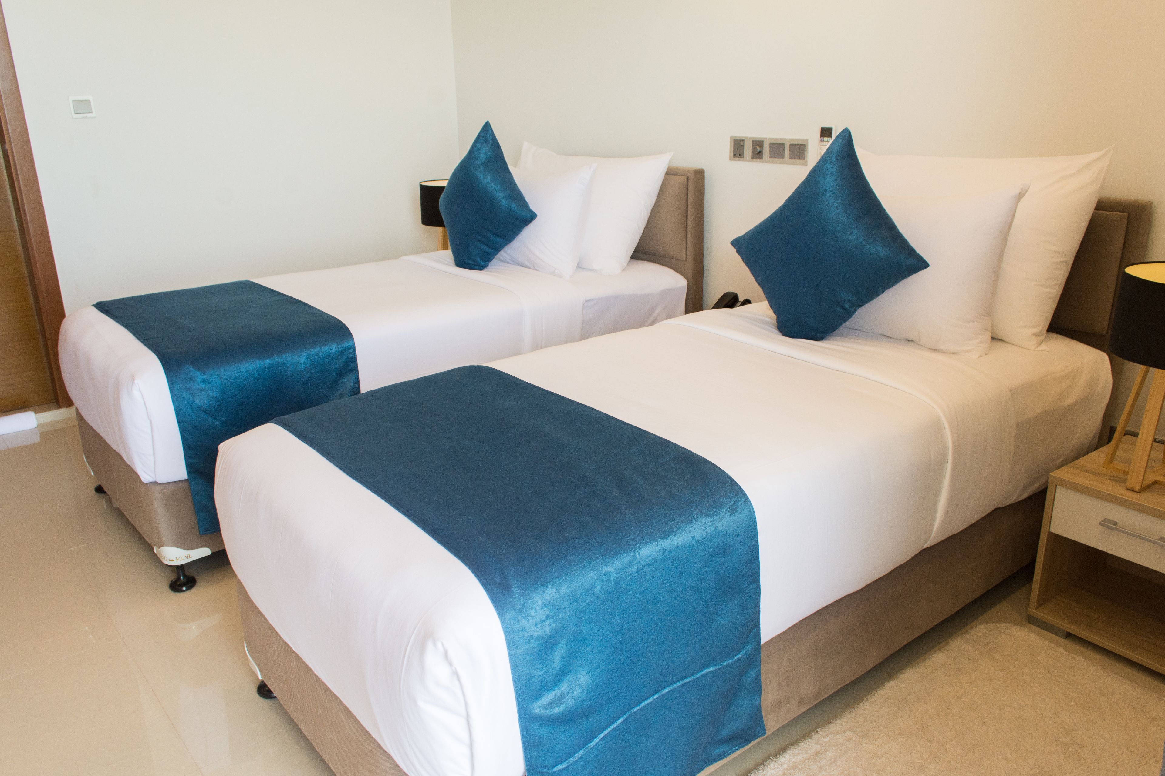Deux lits simples avec du linge de lit blanc et des têtes de lit bleues, une table de chevet en bois avec une lampe, et un mur blanc avec une prise électrique.