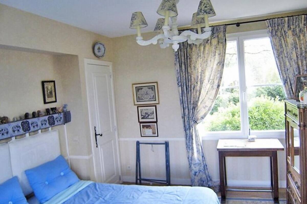 A bedroom with a bed, a small table, a dresser, a window with curtains, and wall-mounted pictures.
