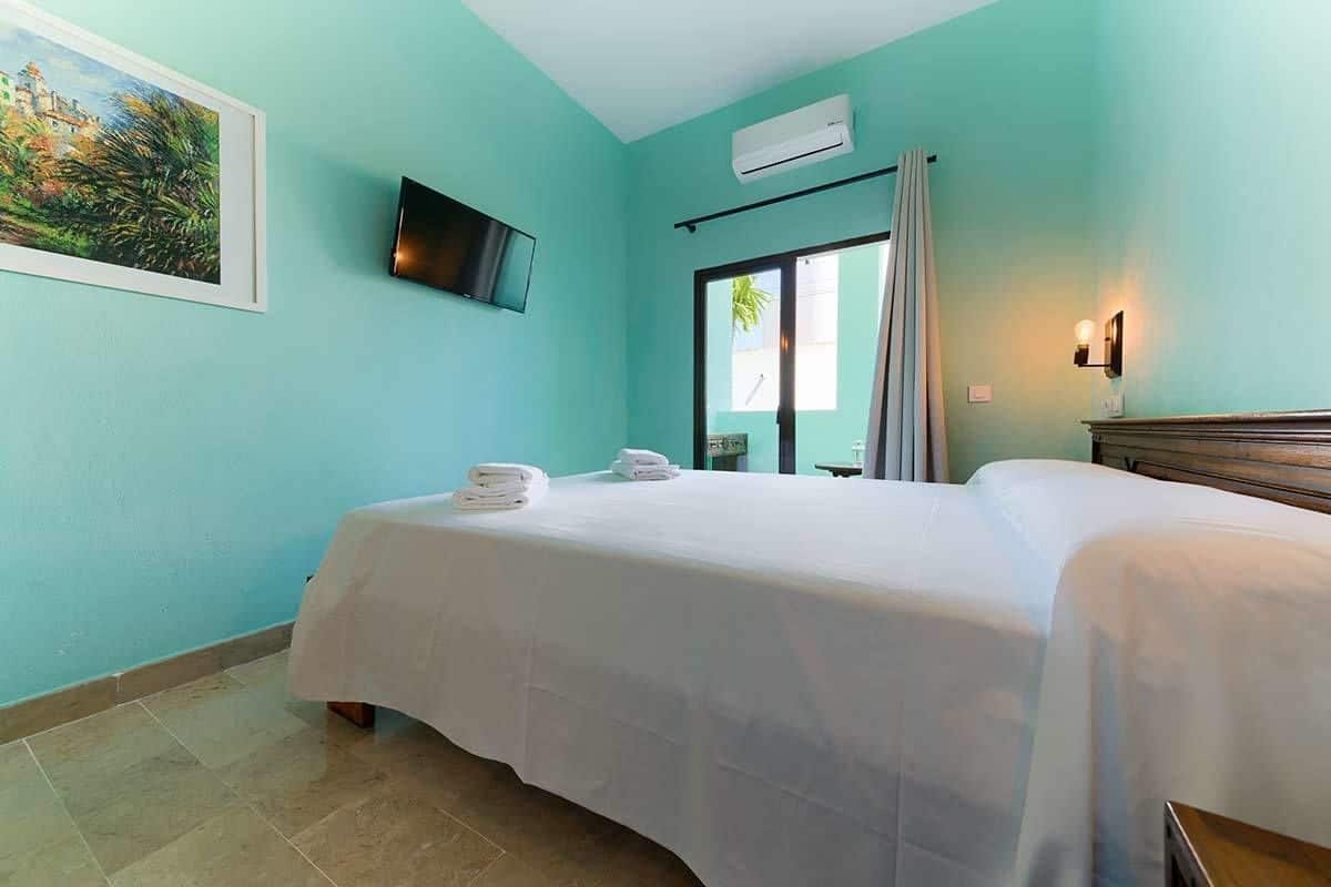 Una habitación con paredes color turquesa, una cama con sábanas blancas, un televisor montado y una ventana con cortinas.