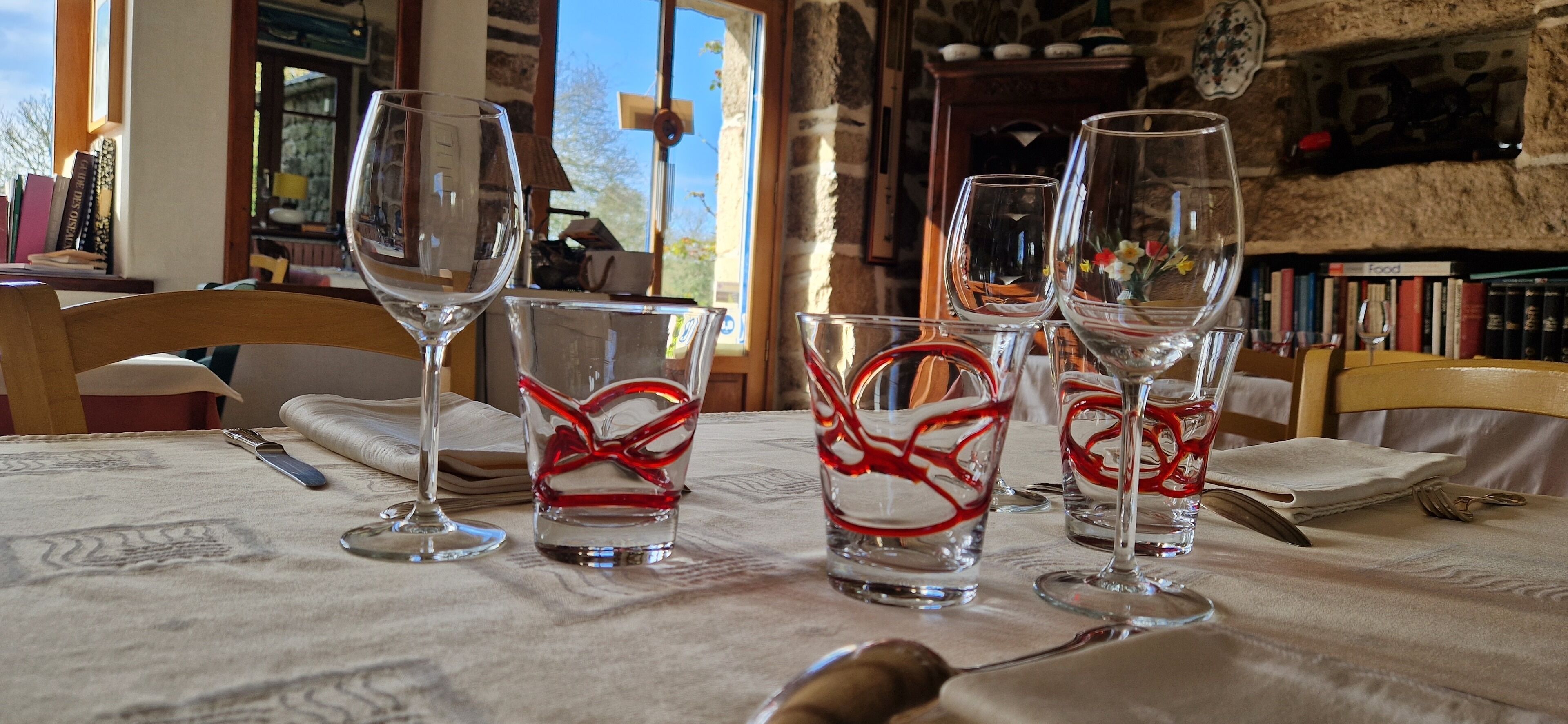 Une table à manger, agrémentée de verres à vin et de couverts, est placée dans une pièce dotée d’étagères et d’un mur en pierre.