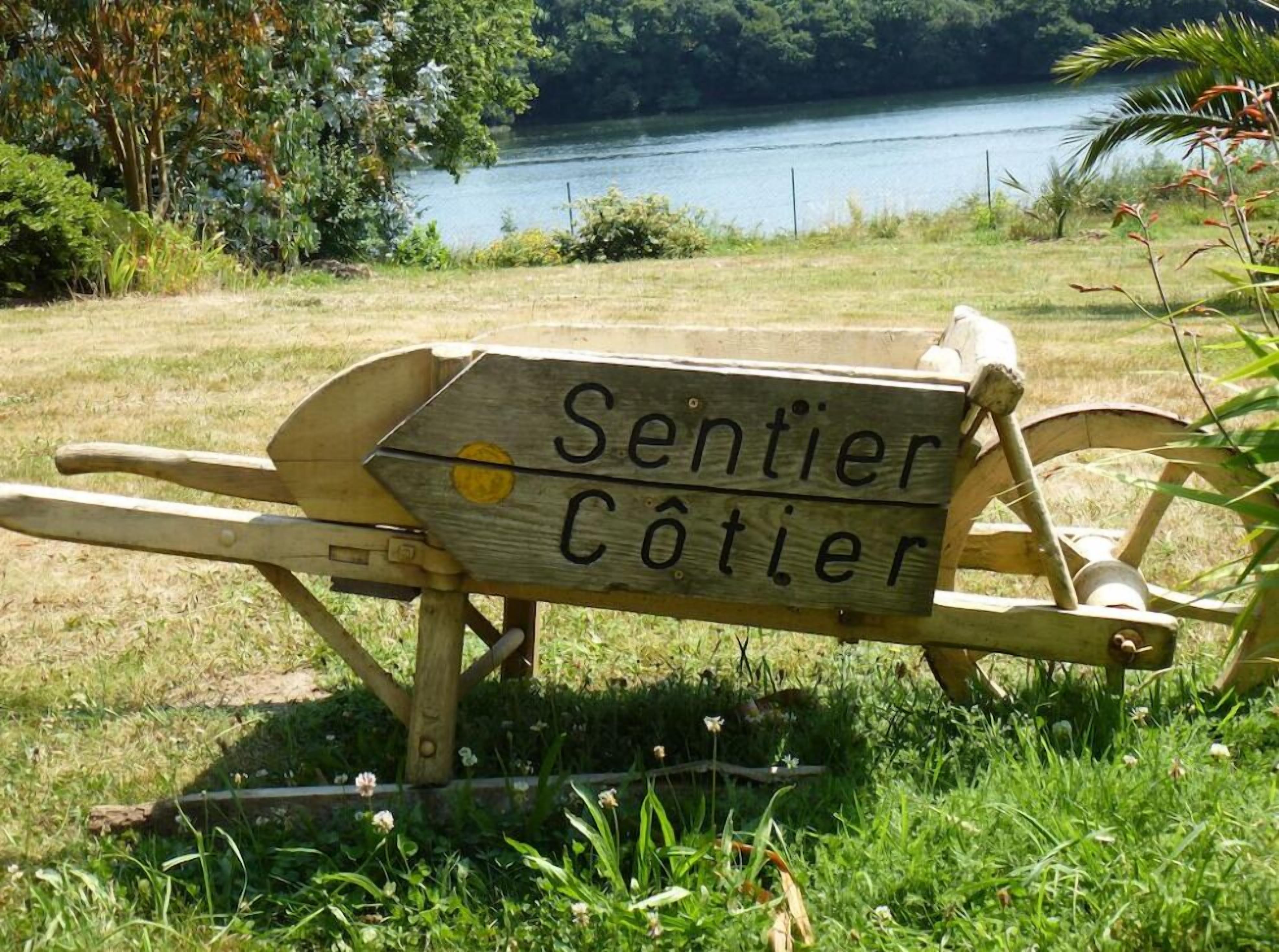 A wooden sign with the text "Sentier Côtier" on a bench in a grassy area with a lake in the background.