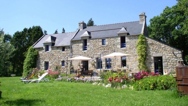 Un bâtiment en pierre avec un toit en pente, de nombreuses fenêtres et un espace extérieur aménagé avec des tables et des parasols.