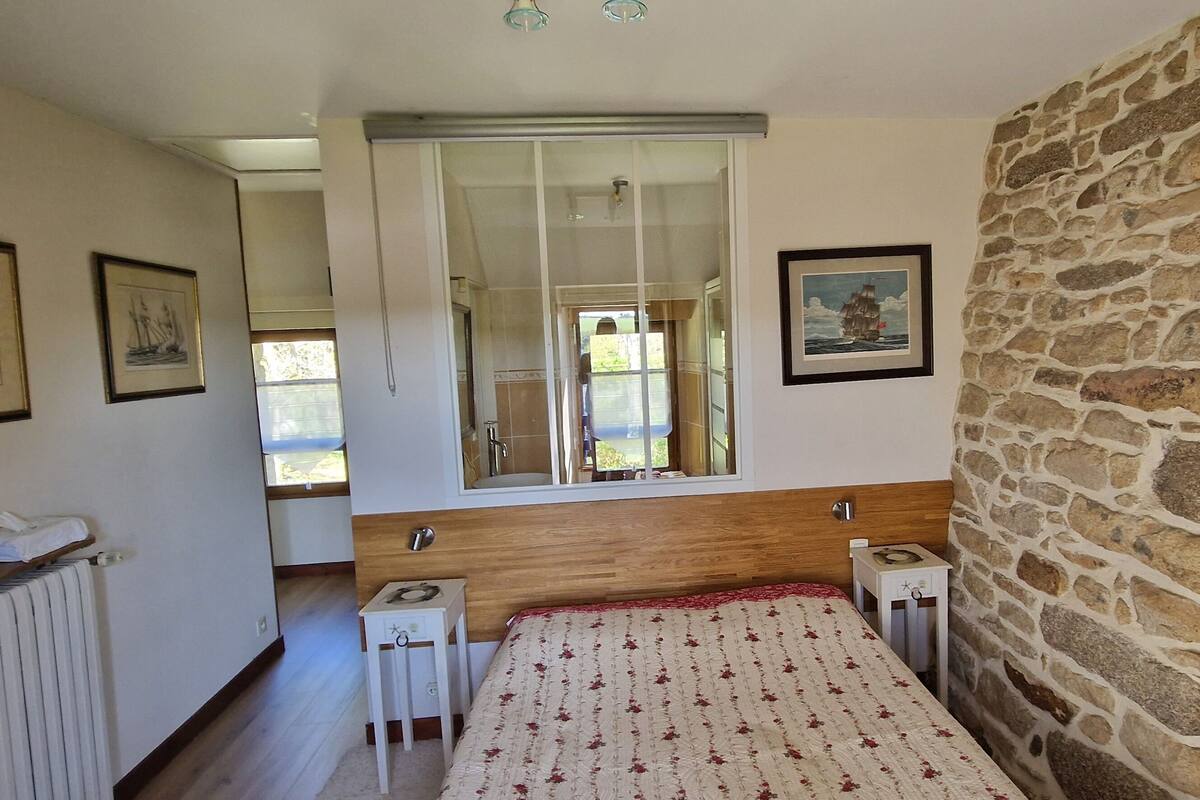 A bedroom with a wooden headboard, a bed with a floral bedspread, a stone wall, and a window with a view of the outdoors.