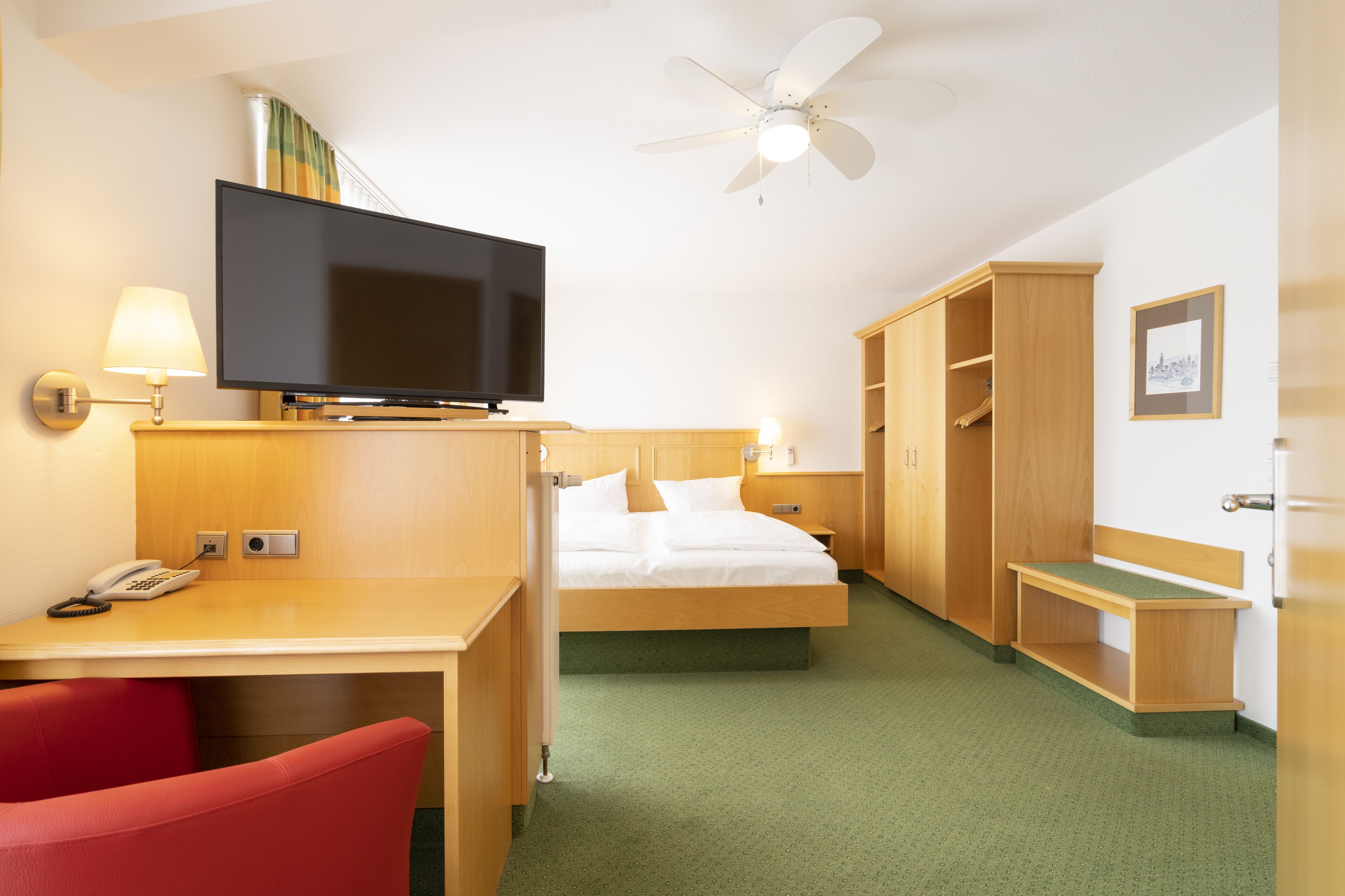 A hotel room with a bed, a desk with a telephone, a television, a wardrobe, and a ceiling fan.
