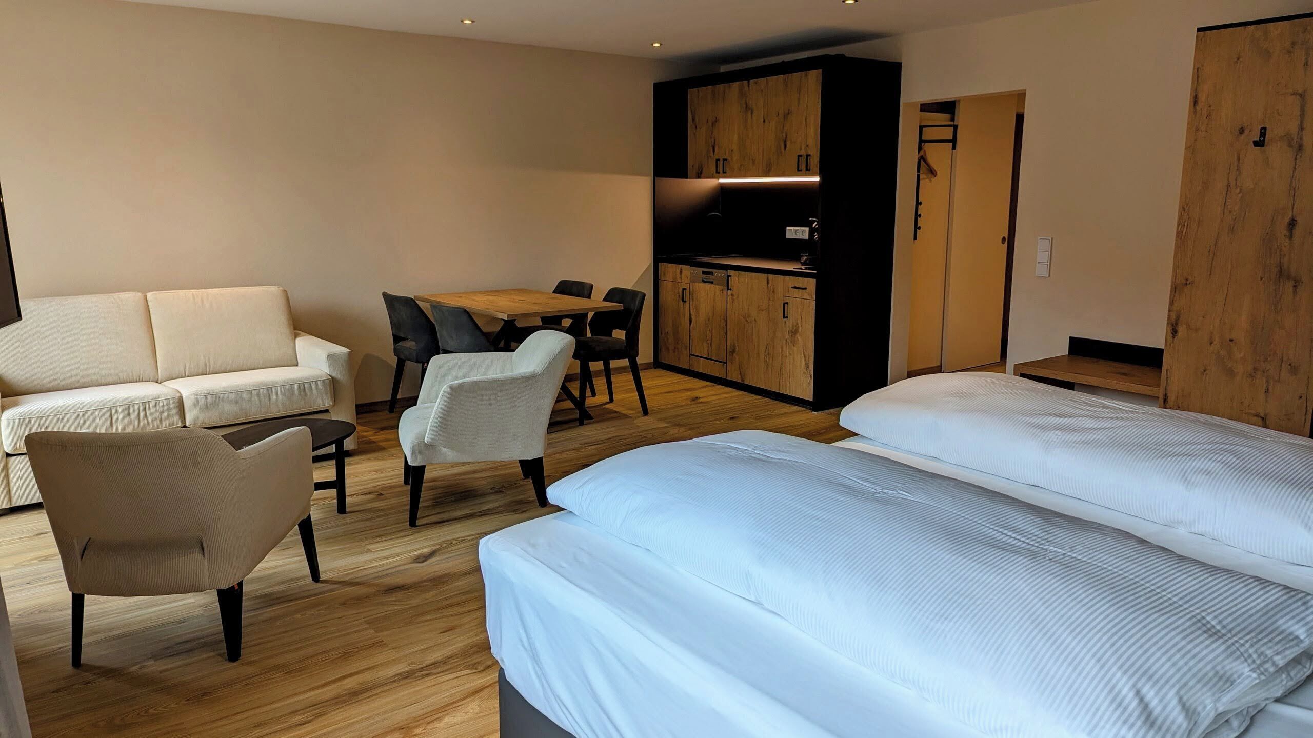 A modern room with a sofa, a dining table, two chairs, a kitchenette, and a bed.