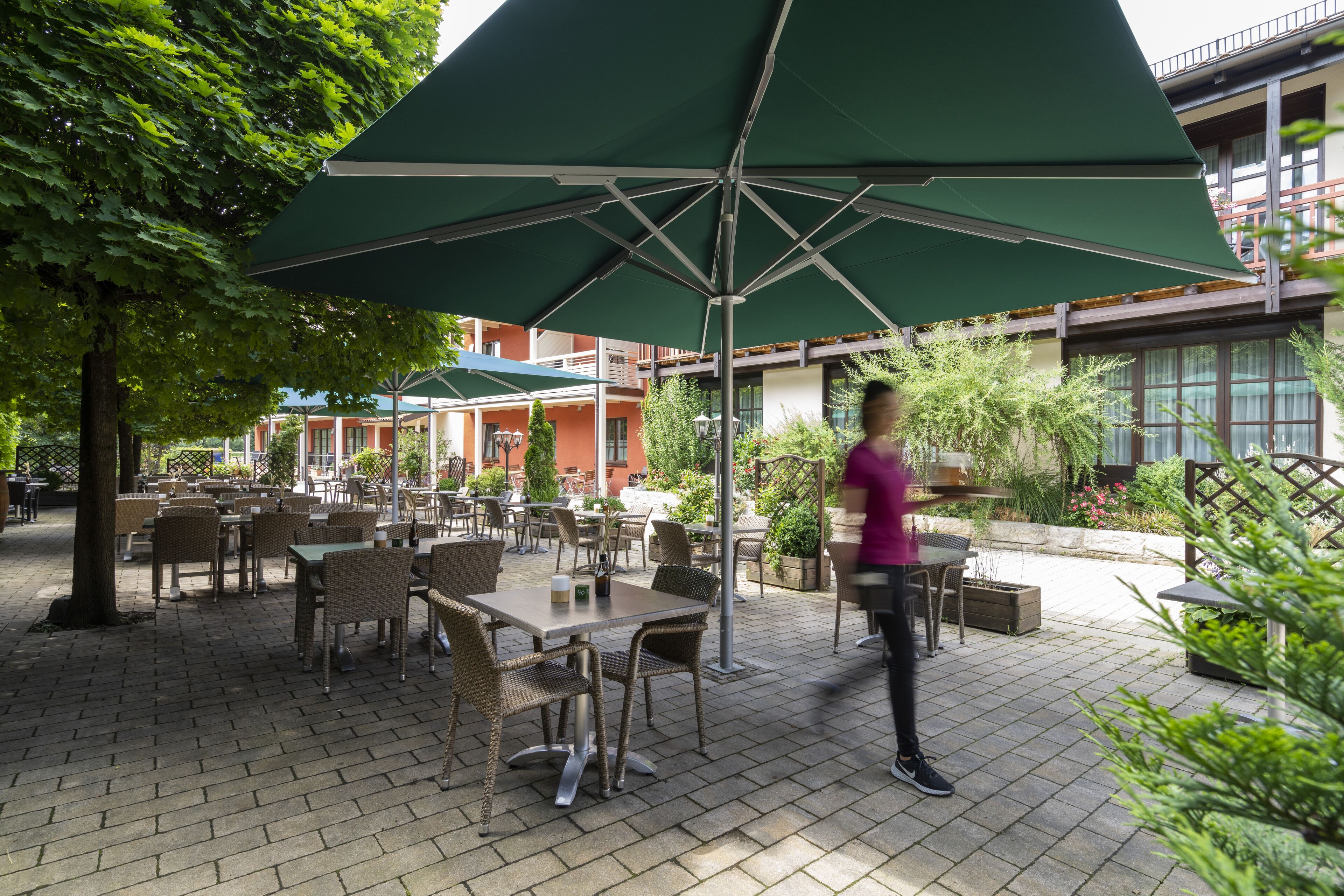 An outdoor patio with tables and chairs under green umbrellas, surrounded by plants and a building in the background.